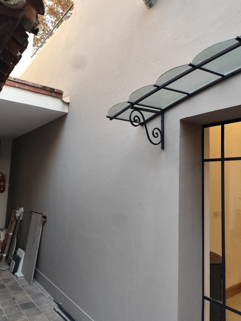 Pared terminada y pintada en gris claro, con un alero de hierro forjado y vidrio sobre una puerta, en un patio.