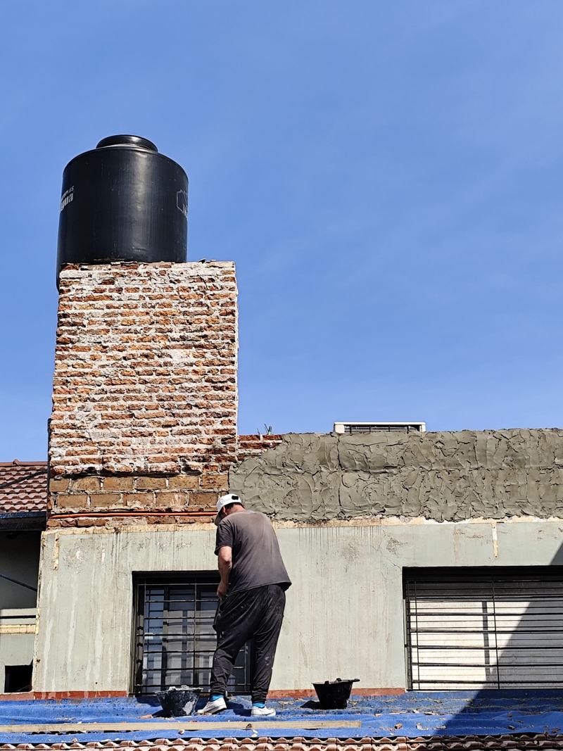 Terraza en obra: un trabajador aplica revoque grueso sobre una pared de ladrillo, junto a una torre de ladrillo con un tanque de agua negro. El techo de tejas está cubierto con una lona azul.