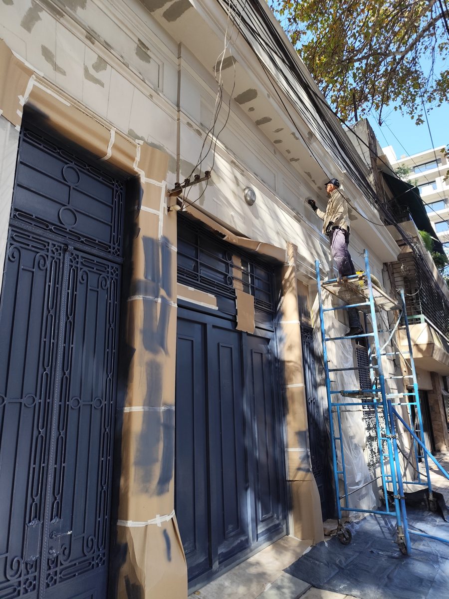 Pintura de fachada con puertas y rejas protegidas con papel y un operario trabajando sobre andamio.